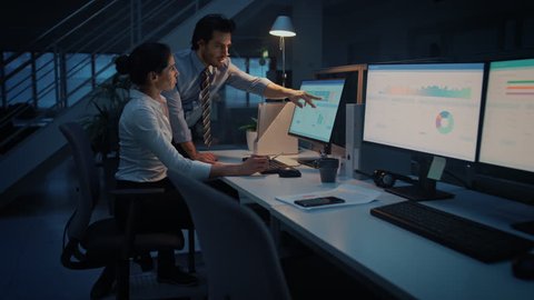 Late at Night In Modern Office: Businessman and Businesswoman Work on Desktop Computer, Having Discussion, Finding Problem Solution, Finishing Important Project. Successful Responsible Office Workers