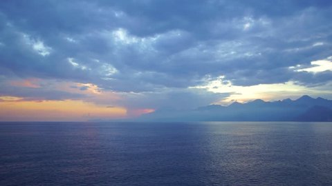 Aerial top view of peaceful calm dark blue sea water surface, cloudy sunset or sunrise sky and silhouettes of mountains. Real time 4k video footage.