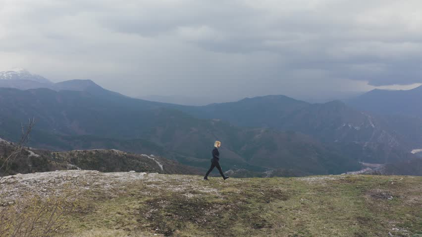 Aerial shot of a girl standing at the top of a mountain with lake in the background. Cloudy and cold weather. 4k