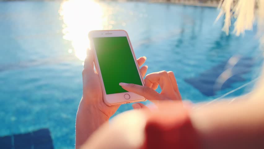 Barcelona, Spain - March 11 2019: Close up of girl using mobile phone green screen while relaxing near the swimming pool. Hands holding smartphone chrome key, tapping modern display smart phone.