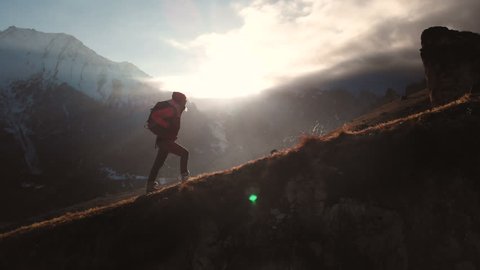 Aerial view of epic shot of a girl walking on the edge of the mountain as a silhouette in a beautiful sunset. Silhouette of a girl in a hat with a backpack climbing uphill