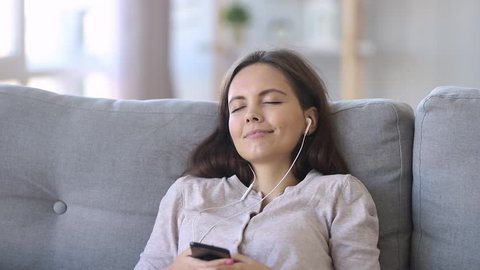 Calm happy young woman in headphones chilling sitting on sofa with eyes closed listening to favorite music holding phone using mobile online player app enjoy peaceful mood wearing earphones at home
