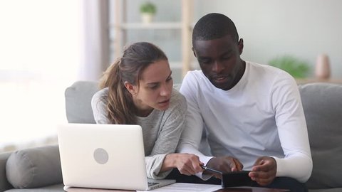 Serious mixed ethnicity young couple discuss pay domestic bills on laptop at home, interracial family planning expenses using calculator computer app managing budget money calculating finances taxes