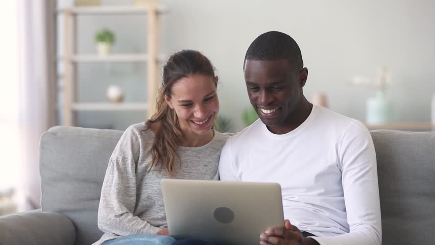 Happy mixed ethnicity young couple using laptop for ecommerce doing online shopping together watching video movie sitting on sofa, smiling african man and caucasian woman looking at computer at home | Shutterstock HD Video #1027488392