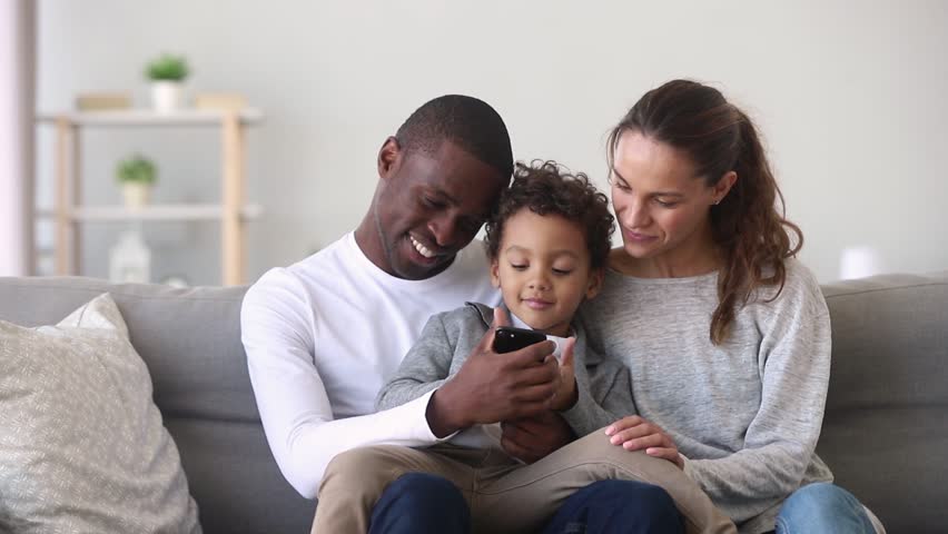 Happy mixed ethnicity family little african dad kid son and caucasian mom holding phone having fun with smartphone, mother father with child boy looking at cellphone enjoy using mobile apps at home Royalty-Free Stock Footage #1027488446