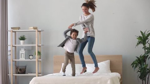 Happy family african american mixed race kid boy and caucasian mom baby sitter holding hands jumping on bed, young mother having fun laughing playing funny active game with cute child son in bedroom