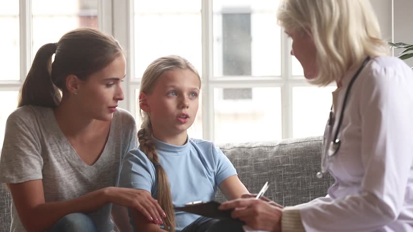 Caring old female doctor pediatrician talk to school child girl make notes in clipboard listen to little patient sit on sofa with mother tell complaints at medical checkup appointment consultation | Shutterstock HD Video #1027488497