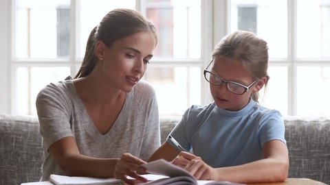 Caring mom babysitter teacher helping kid daughter with homework, parent mother teaching child read book and cute school girl learning writing in textbook studying at home, family education together