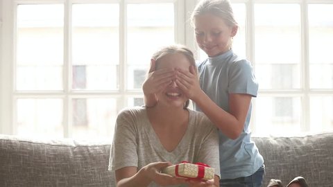 Cute kid daughter covering mum eyes making surprise to happy young mom, child girl hugging excited mommy congratulating with mothers day concept presenting gift box embracing sitting on sofa at home 