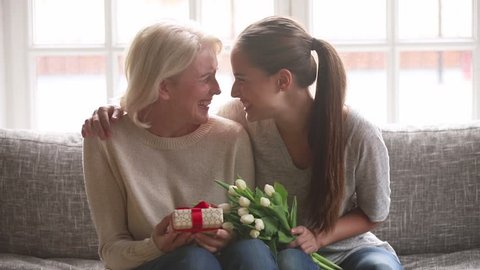 Loving adult daughter presenting spring flowers and gift box to happy old mother laughing bonding embracing, young woman congratulating hugging senior mom looking at camera on mothers day at home
