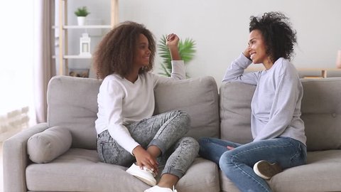 Happy best friends african mother and teen daughter talking sitting on sofa at home, friendly black family loving mum with teenager girl chatting on couch share secrets having fun trust conversation