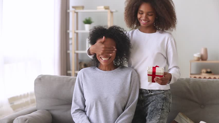 Mothers day present concept, african american teen daughter make surprise congratulating happy excited black mommy closing mum eyes embracing mixed race mom giving gift box hugging on couch at home Royalty-Free Stock Footage #1027488584