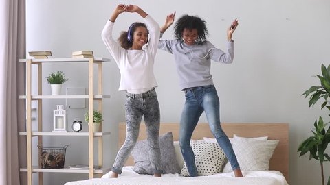 Happy carefree family black mom and teen daughter wearing headphones dancing on bed listening to music on phone, funny funky african american mother sister and teenager girl having fun in bedroom