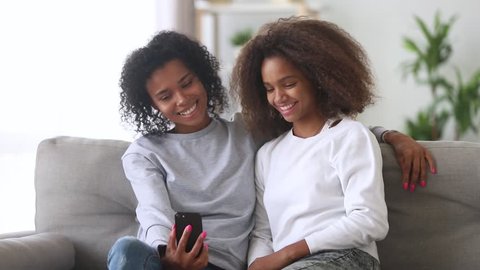 Happy african american family mother and teen girl using mobile app taking selfie looking at smartphone, smiling black mom with daughter having fun make phone snapshot self portrait sitting on sofa