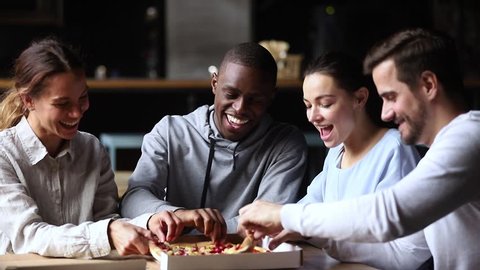 Multicultural happy friends talking laughing sharing takeaway pizza meal together in cafe indoors, smiling diverse young mates students having fun eating food together at meeting sitting at table