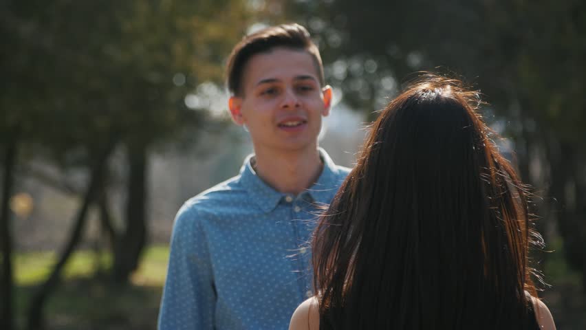 Young boy came to his girlfriend, kiss her and showed a camera behing her. Young positive couple in the park.