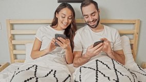 Pleased lovely couple using smartphones while lying together on bed in bedroom