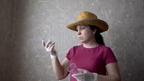 Woman examines plastic cups. On his head a straw hat. Burgundy t-shirt. Gray Wallpaper on the wall. Home-room. A doctor, a vet, a gardener, a villager. 4K video.