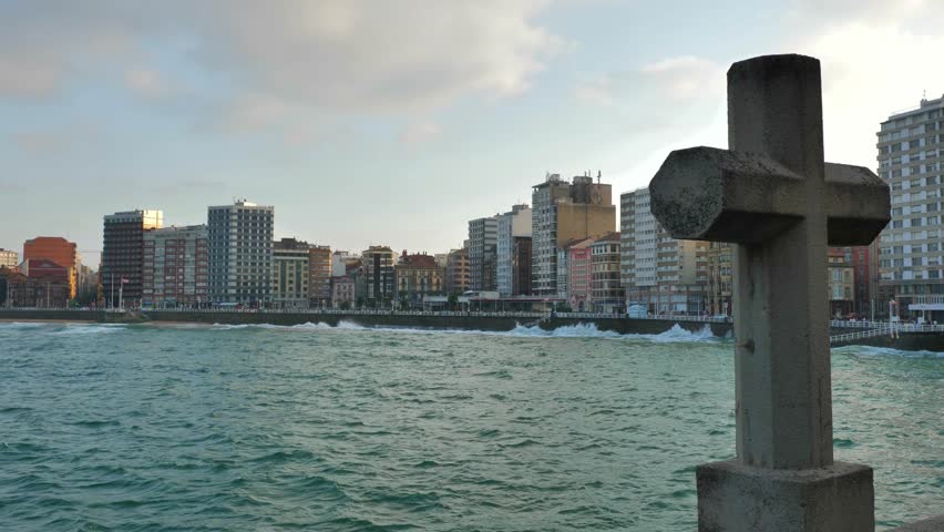 The skyline of the city of Gijon (Asturias, Spain) with a stone cross on the right of the frame. High buildings facing the sea, waves moving the water. Clouds creating an even lighting.