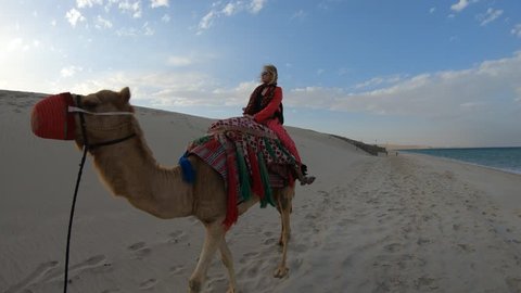 Inland sea is a major tourist destination for Qatar. Freedom woman riding camel on beach at Khor al Udaid in Persian Gulf. Caucasian blonde tourist enjoys camel ride in Middle East, Arabian Peninsula.