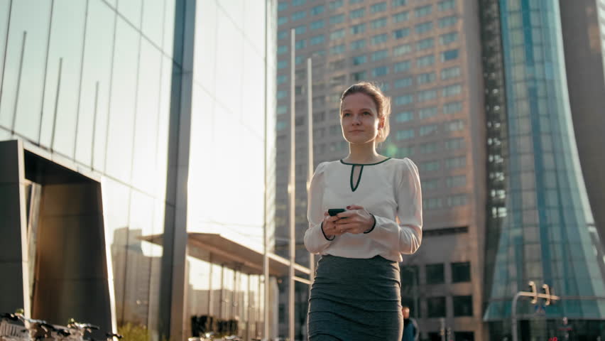 Adult Caucasian Confident Young Business Woman with Hair Bun is Using Smartphone App and Walking Outside near Modern Office Building. Medium Long Low Angle 4K Slow Motion Corporate Tracking Shot
