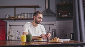 Smiling handsome bearded man having breakfast and using smartphone while sitting by the table at home