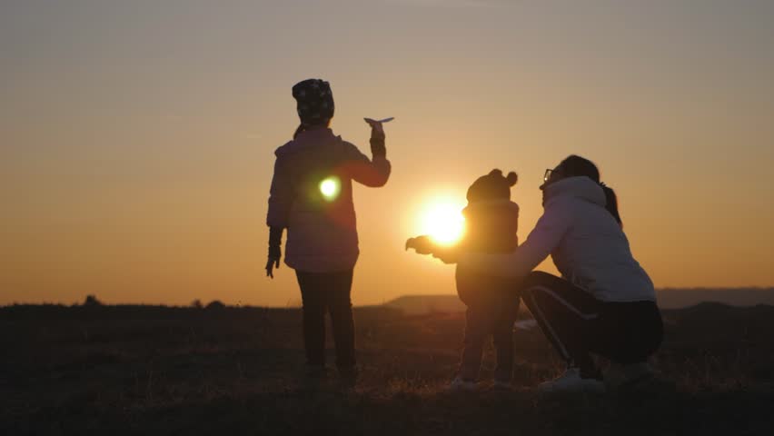 Little girl playing with a paper airplane, mom and little brother are looking at her. Silhouette of mother and two children at sunset. Concept of dreams and travel. | Shutterstock HD Video #1027551563