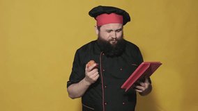 cheerful chef with a beard in a black suit on a yellow background, uses a tablet, talking on video calls, laughs and eats a tomato