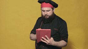 cheerful chef with a beard in a black suit on a yellow background, uses a tablet, talking on video calls, laughs and eats a tomato