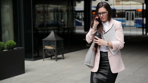 A Young Stressful Business Woman in the Glasses Goes Through the City With Documents and Talks on the Phone. Coffee Time.