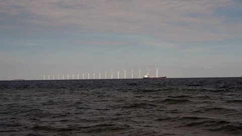 Wind turbines in motion at sea with blue sky and open water, with a kite surfer, off the coast of Denmark, April 2019