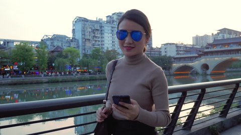 Lovely and young Asian woman is walking alongside the Funan River of Jiuyan Bridege in Chengdu, as smiling in front of the camera with mobile phones in her hand.