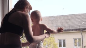 A beautiful young woman and her adorable little son are playing by the window and mother shows to the baby boy something behind the window in slow motion middle shot 4K indoor video