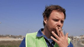 a worker with a beard and mustache smokes a cigarette and puts on a yellow helmet. 4k video