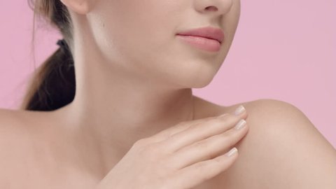 Close-up beauty portrait of young Caucasian woman with smooth and healthy skin strokes her skin at collarbone area against light pink background | Skincare concept