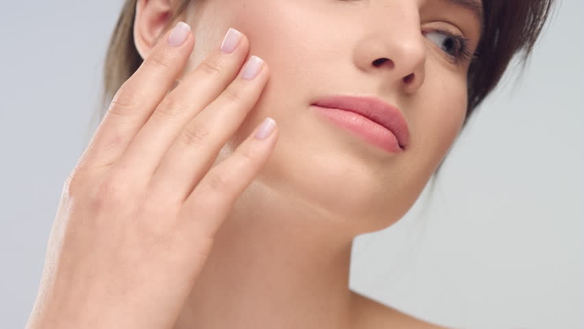 Close-up beauty portrait of young woman with smooth healthy skin, she gently touches her face with her fingers on light grey background and smiles | Skincare concept