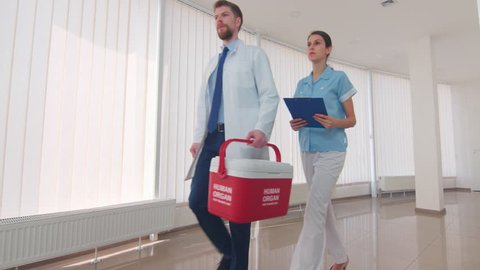 Medical courier with an assistant go down the clinic hall with red human organ trafficking container | Human organ transplantation concept
