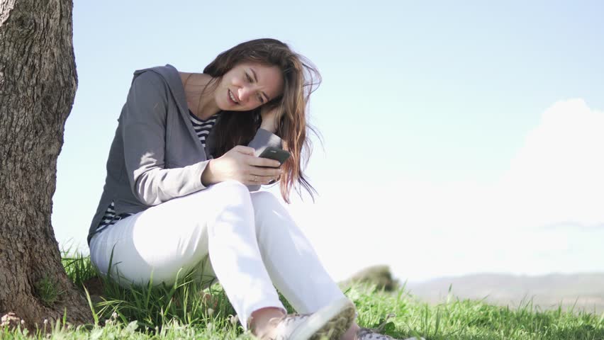 A young woman plays on a mobile phone. She sits under a tree in a field by the sea and smiles. The girl writes a message on the phone in nature