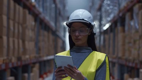 Beautiful young woman worker in warehouse of shopping center. Girl looking for goods with a tablet is checking inventory levels in a warehouse. Logistics concept