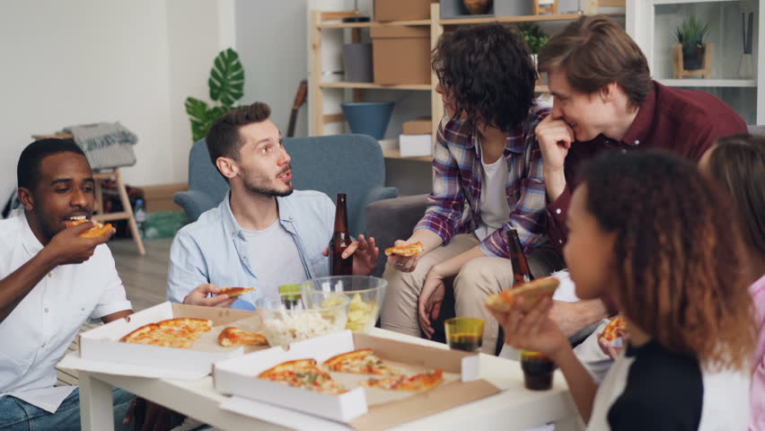 Young people friends are eating pizza, chatting and laughing at funny party in modern apartment.Men and women are holding food and drinks bottles and glasses. | Shutterstock HD Video #1027624217