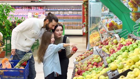 Young parents with a little daughter choose fresh apples in a large supermarket. Happy family buying groceries at the vegetable store. Slow motion.