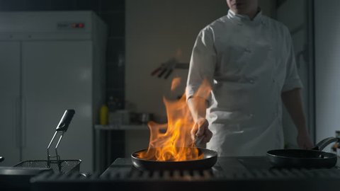Chef fires up the flambe on a hot pan at the kitchen in slow motion, big open fire in the kitchen, pan on fire, cooking on the open flame, 4k UHD 60p Prores HQ 422: film stockowy