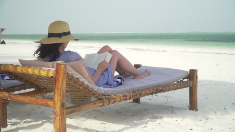 Young woman is reading book on white sand beach by ocean. Adult caucasian girl in straw hat and sundress is lying on lounger sunbed in shadow on white sandy beach and reading book at bright sunny day.