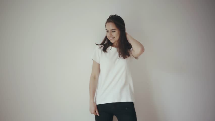 Young beautiful girl posing in front of the camera in white cotton blank t-shirt and black jeans with white isolated wall on the background, mock up of clothes, happy smiling and laughing