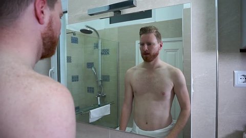 Young Skinny caucasian man trying to flex his muscles in front of the mirror in the bathroom wearing white towel