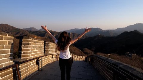 Tourist enjoy Badaling Great Wall of China, standing with arms spread out at high point. Beautiful mountains around, long strip of wall follow hilly terrain. Warm sun light at evening hour