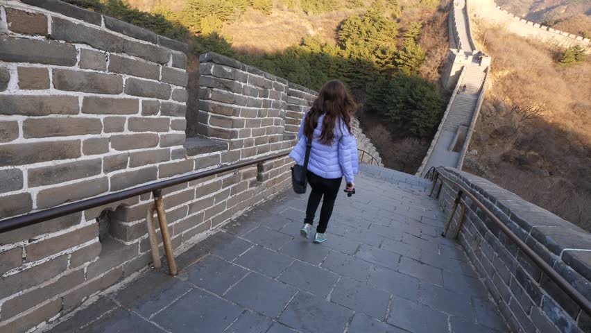 Tourist woman at Great Wall of China, come down at steep slope, very sharp stairway ahead. View from above, fortification follow hill side and continue on bottom