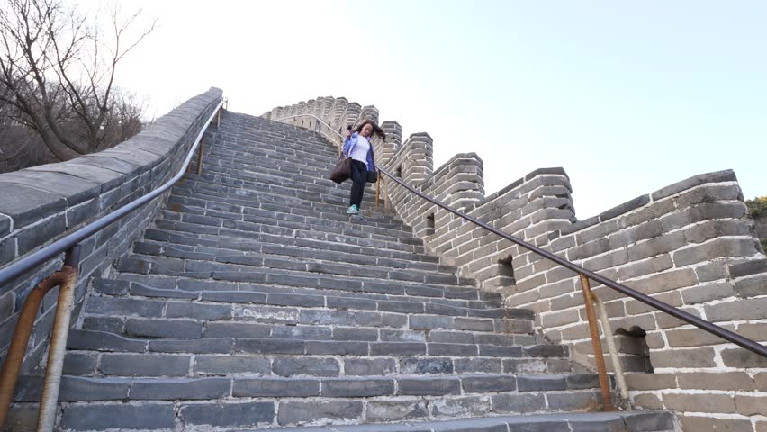 Tourist woman walk down on steep stone stairs, visit Great Wall of China, Badaling section. Camera looking up to sharp descent. Lady hold small camera and handbag, watch steps while going downstairs