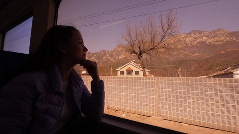 Tourist woman travel by Chinese train S2 to Badaling Great Wall, sit against window, look out. Small settlement and fence along rail track seen outdoors, mountains seen on background