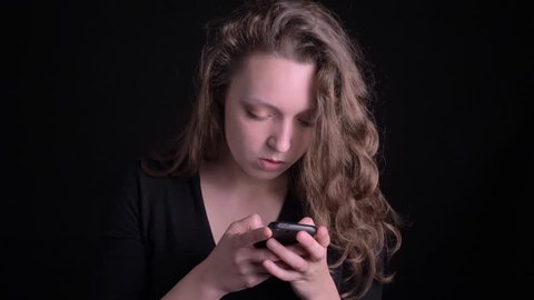 Portrait of young curly-haired girl watching seriously and attentively into smartphone on black background.